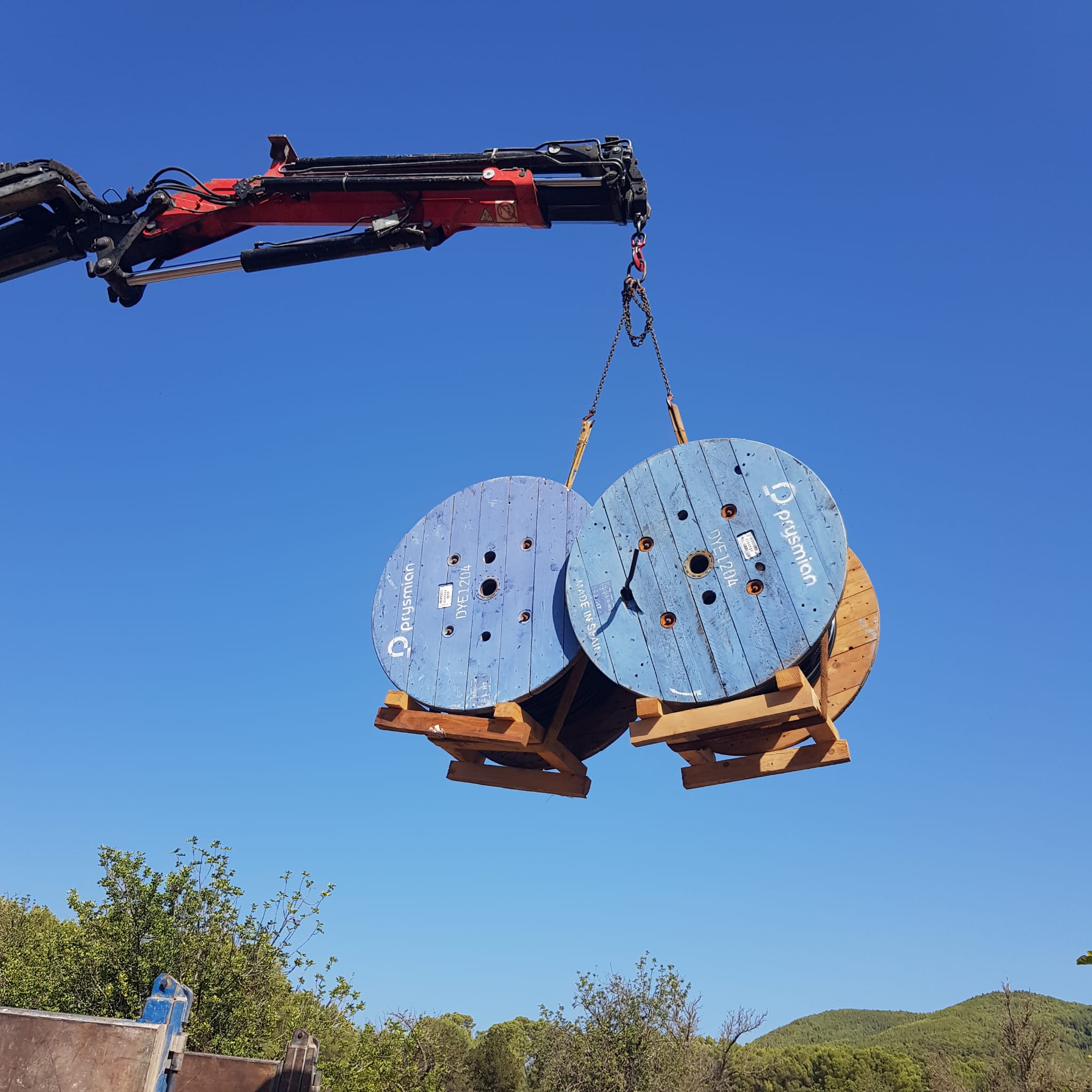 Grúa elevando un palé con dos bobinas de cable bajo el cielo azul